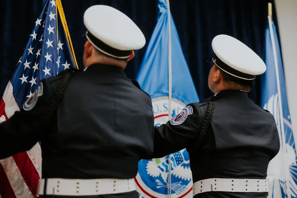 Two U.S. Navy sailors in uniform standing in front of American and military flags during a ceremonial event.