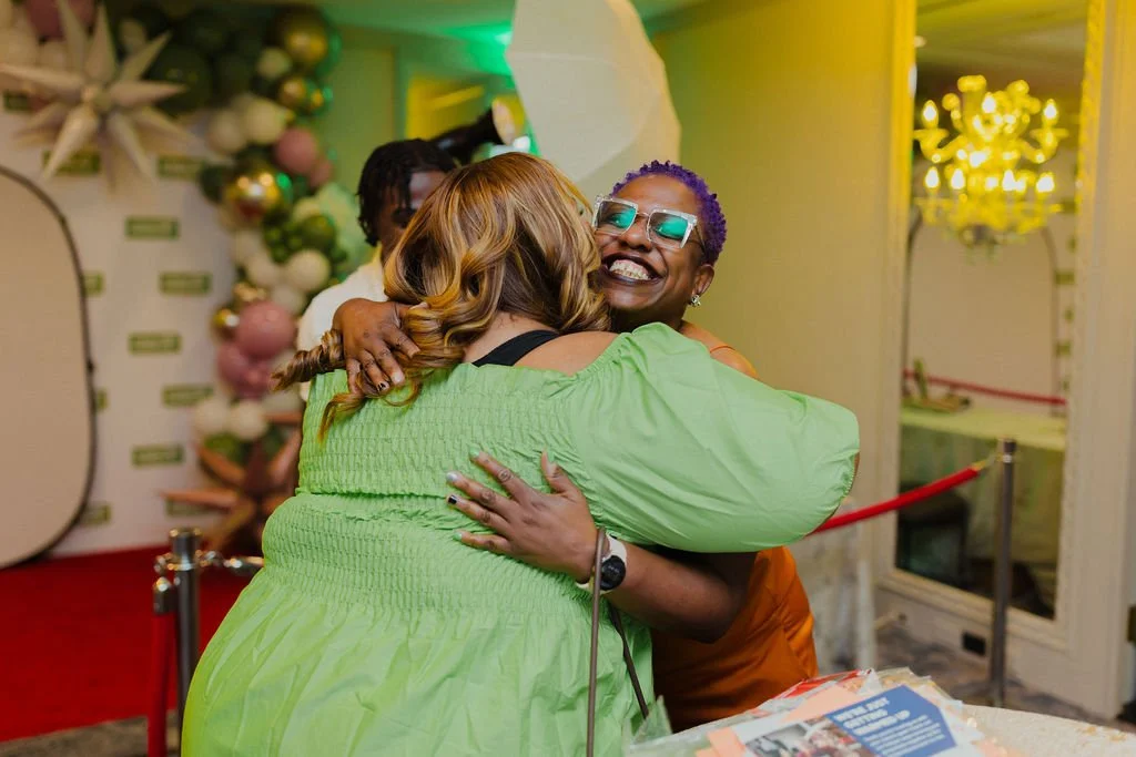 Two women hugging warmly at a festive event. One woman is smiling broadly, wearing sunglasses and an orange top. The other woman, with light brown hair, is wearing a bright green dress. The background features holiday decorations and a chandelier.