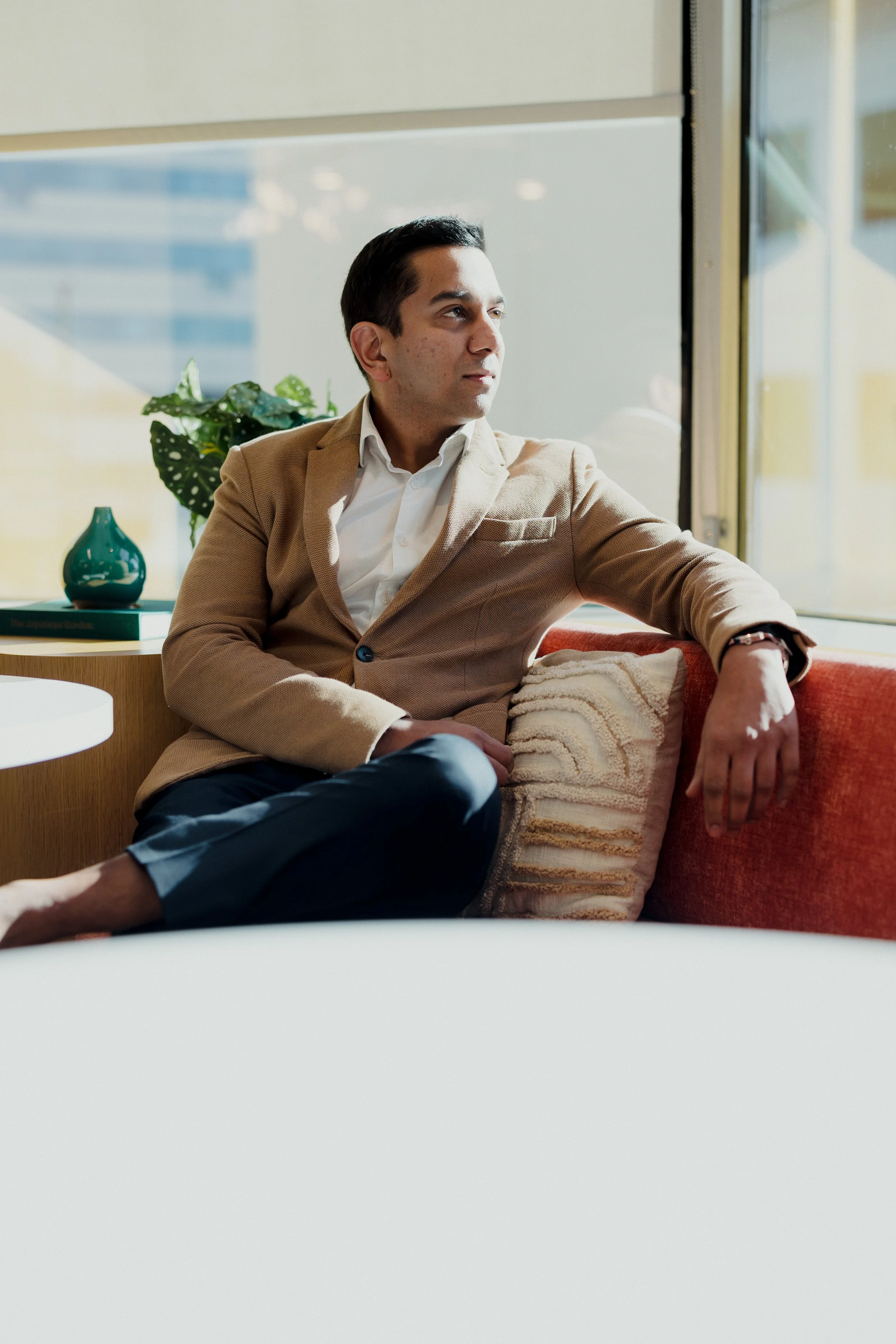 A man in a beige blazer and white shirt sitting on a red sofa near a window, looking out thoughtfully.