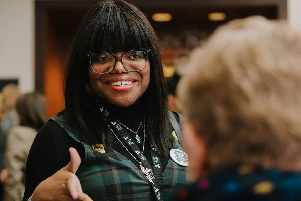 A smiling woman with black hair, glasses, and a black and green plaid vest giving a thumbs-up in an indoor setting.