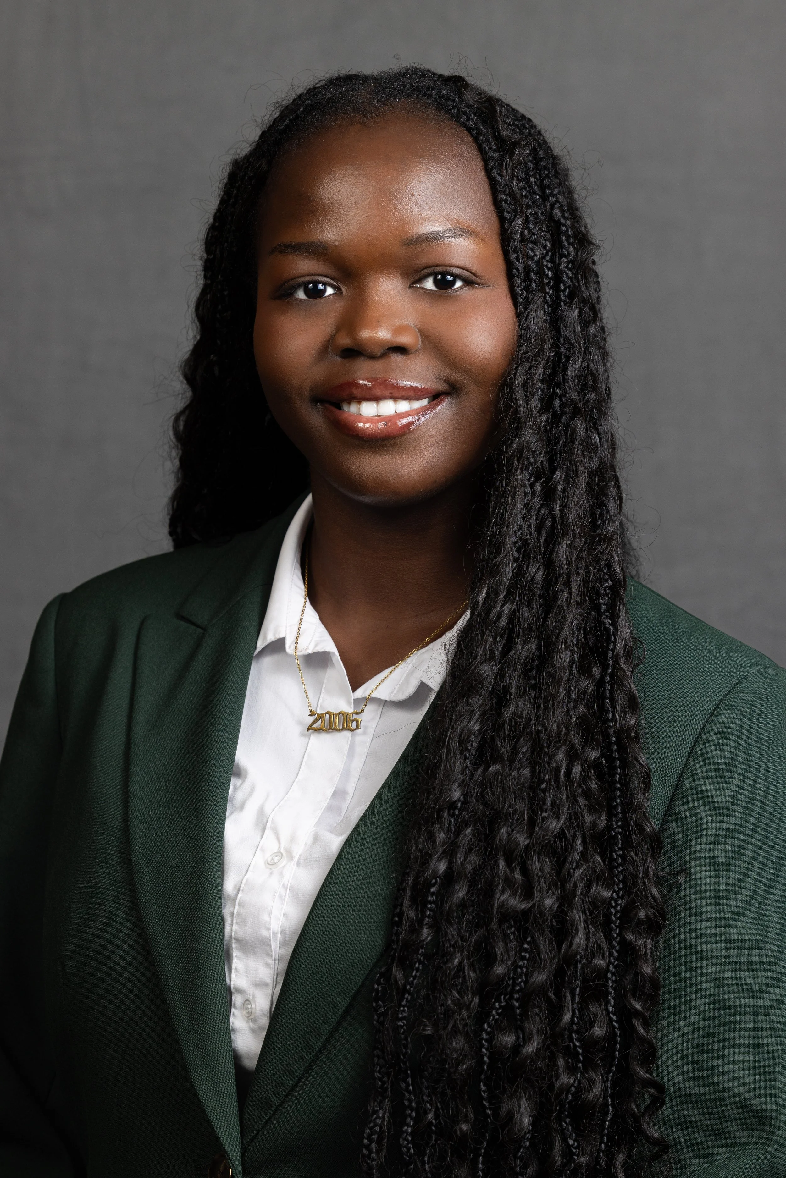 A woman with dark skin, long curly black hair, wearing a white shirt, dark green blazer, and a gold necklace with the year '2015' pendant, smiling against a gray background.
