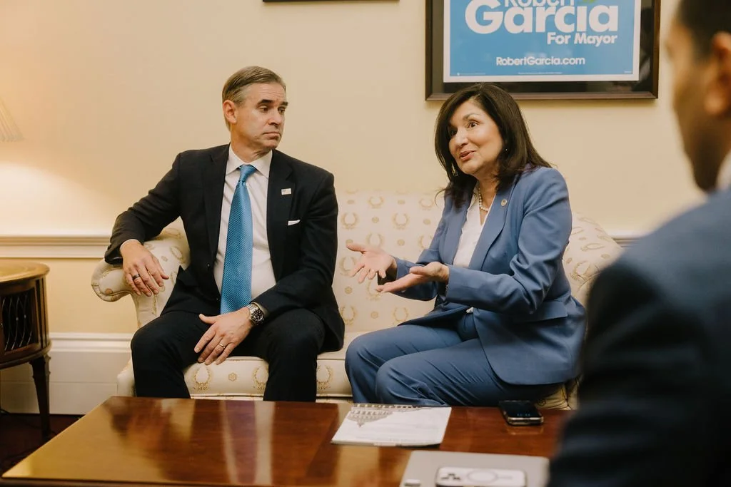 A woman in a blue suit speaking in a meeting room, seated on a sofa with a man in a suit and blue tie listening.