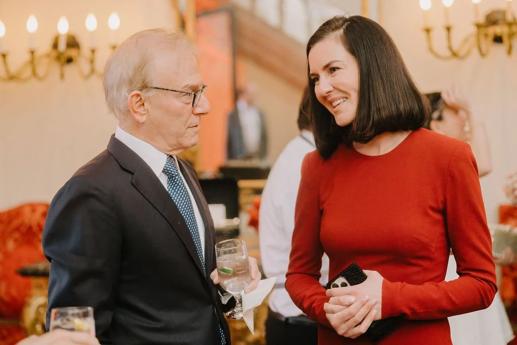 A man in a suit and tie holding a glass of water, talking to a smiling woman in a red dress at a social gathering.