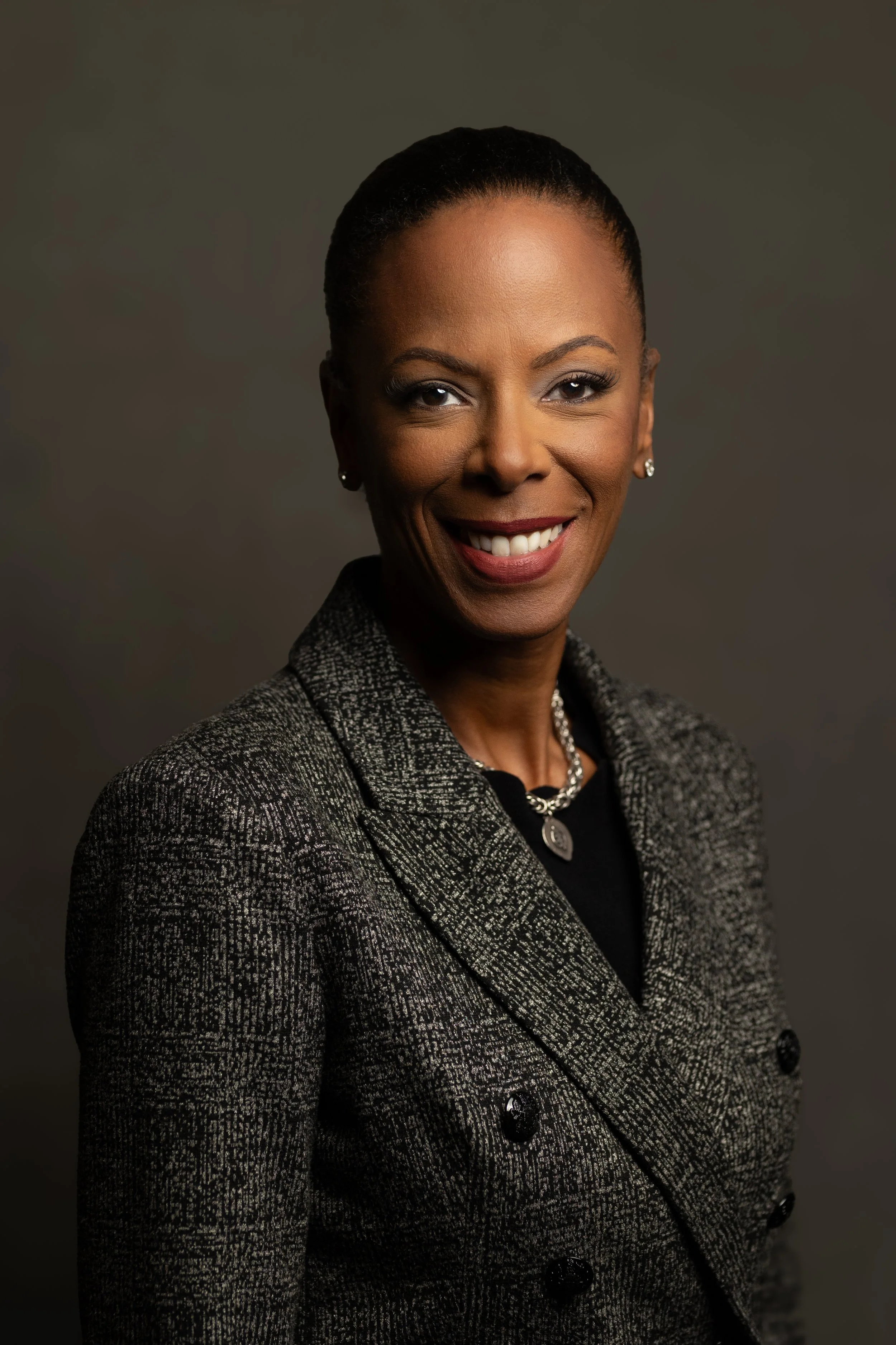 A professional woman with short hair wearing a gray plaid blazer and jewelry, smiling against a dark background.