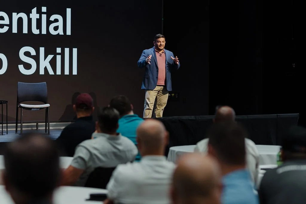 A man in a blue blazer and khaki pants giving a presentation on stage to an audience seated at round tables. The background features large text on a screen.