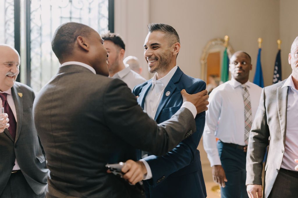 Two men in suits greeting each other with a handshake and smile, in a formal indoor setting with other businesspeople in the background.