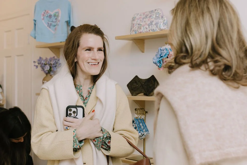 Two women smiling and talking inside a store, with shelves displaying bags, scarves, and decorative items in the background.