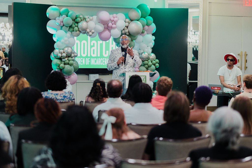 A speaker at a podium addresses an audience during an event called 'NolarCh', focused on children of incarceration. The speaker is an elderly man with gray hair, wearing glasses, a gray patterned jacket, and holding a microphone. Behind him is a deco