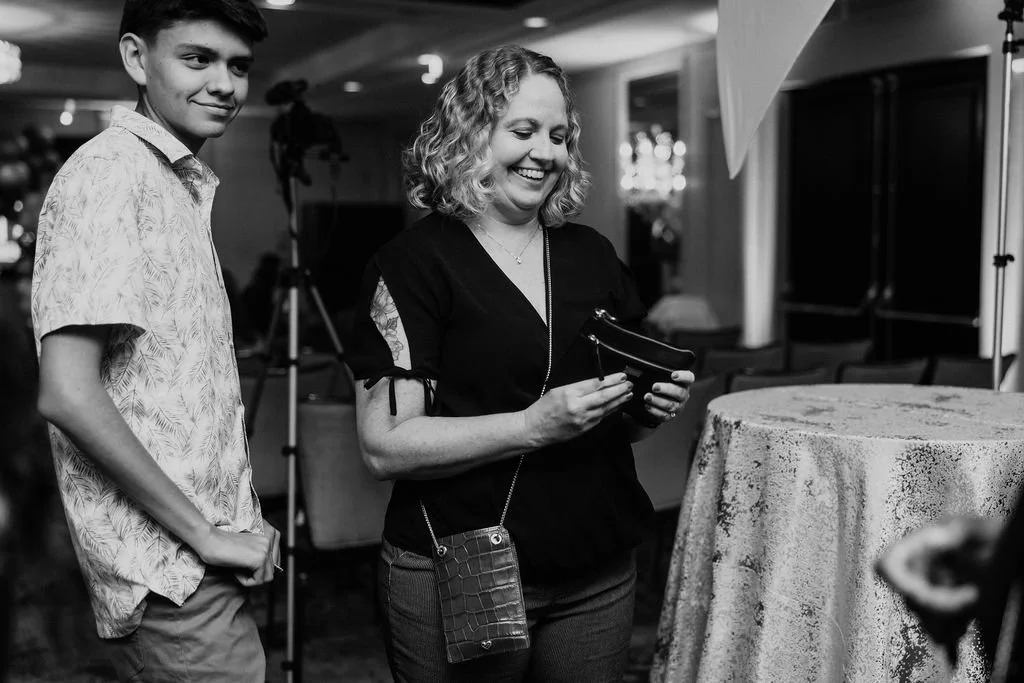 A woman smiling while looking at her phone, standing next to a teenage boy in a patterned shirt in an indoor setting with photography equipment and tables.