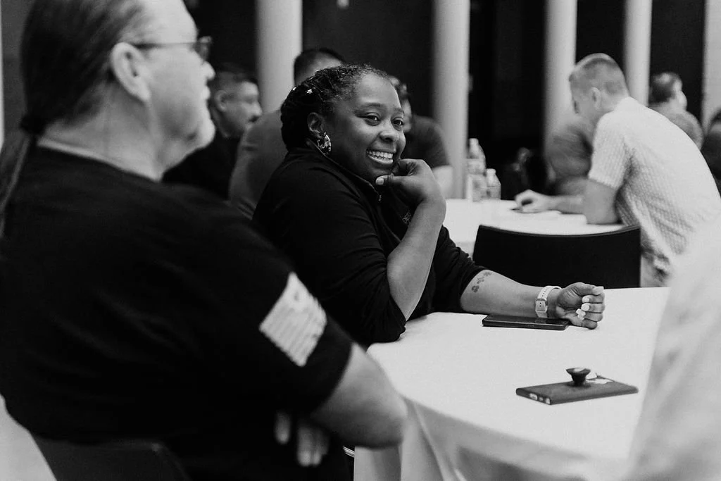 A woman with braided hair smiling and resting her chin on her hand, sitting at a table in a conference room with other people.