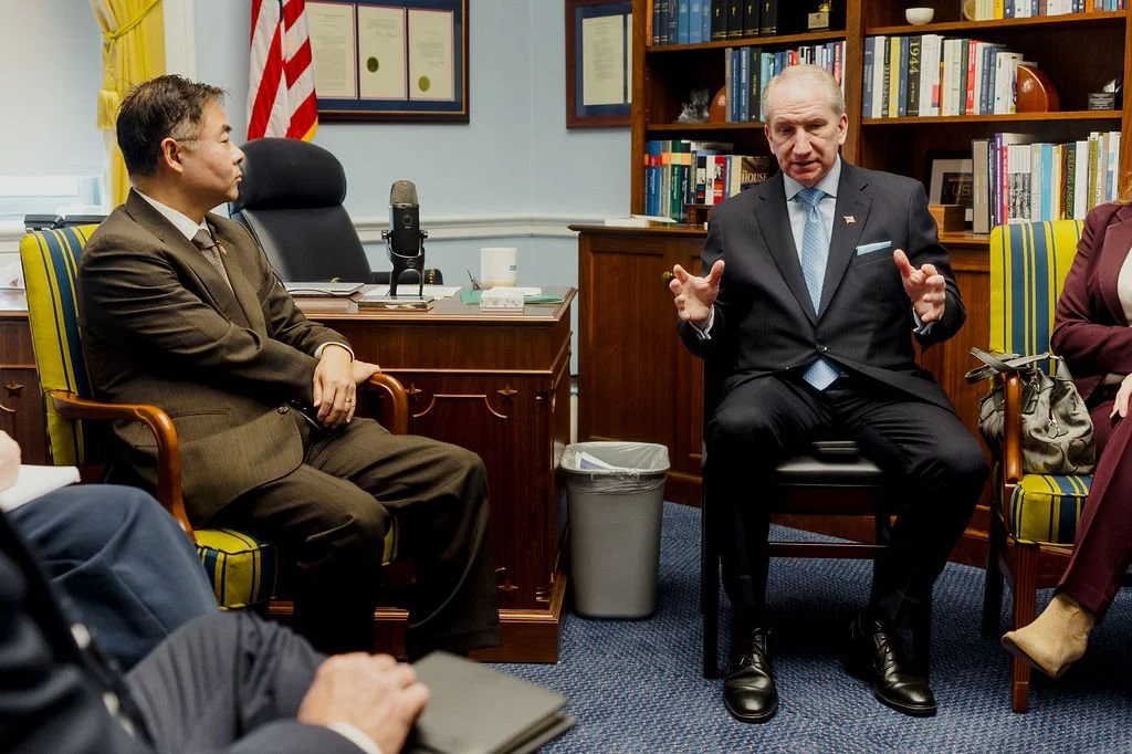 Two men sit and speak in a formal office setting, with bookshelves and framed documents on the wall behind them. One man, on the right, is wearing a dark suit and tie, and the other, on the left, is dressed in a brown suit. They are engaged in conver