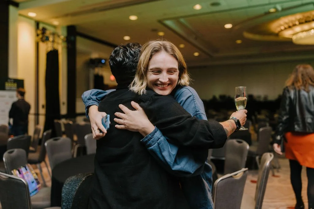 Two people hugging at a celebration or event in a banquet hall, with one holding a glass of champagne.