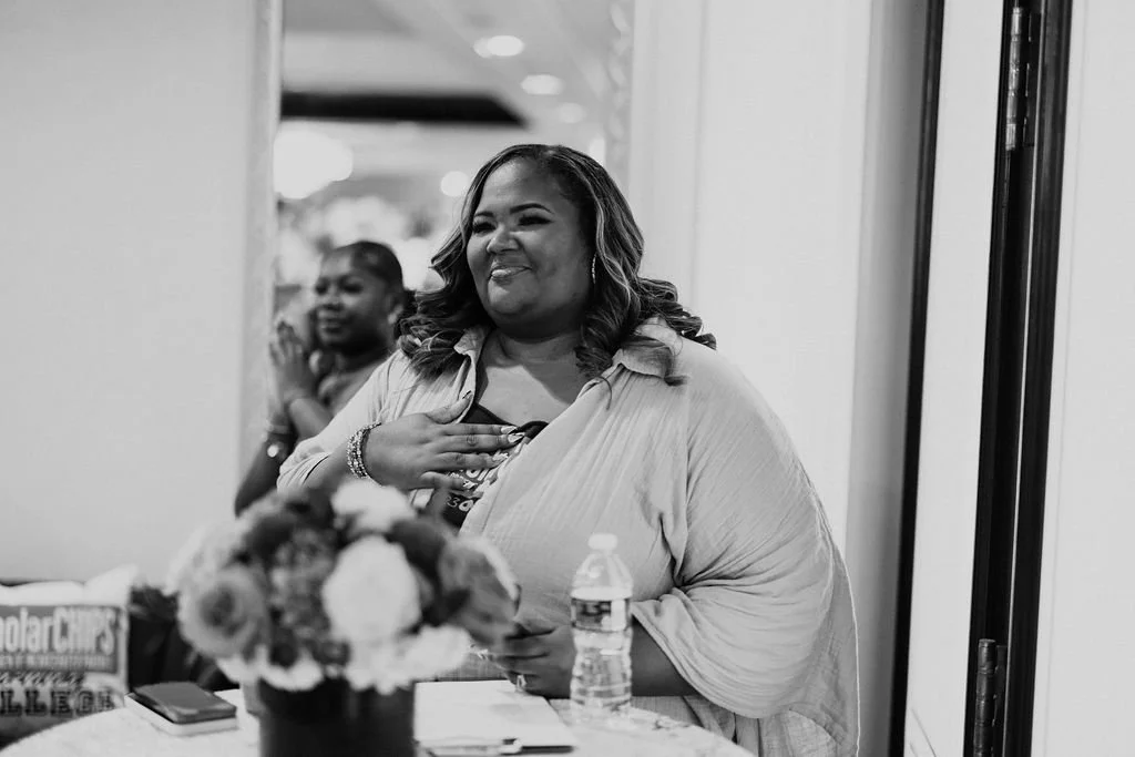 A smiling woman with wavy hair wearing a light-colored jacket, holding a bouquet and a water bottle, standing at a table with a flower arrangement during a celebration or event, with another woman in the background clapping.