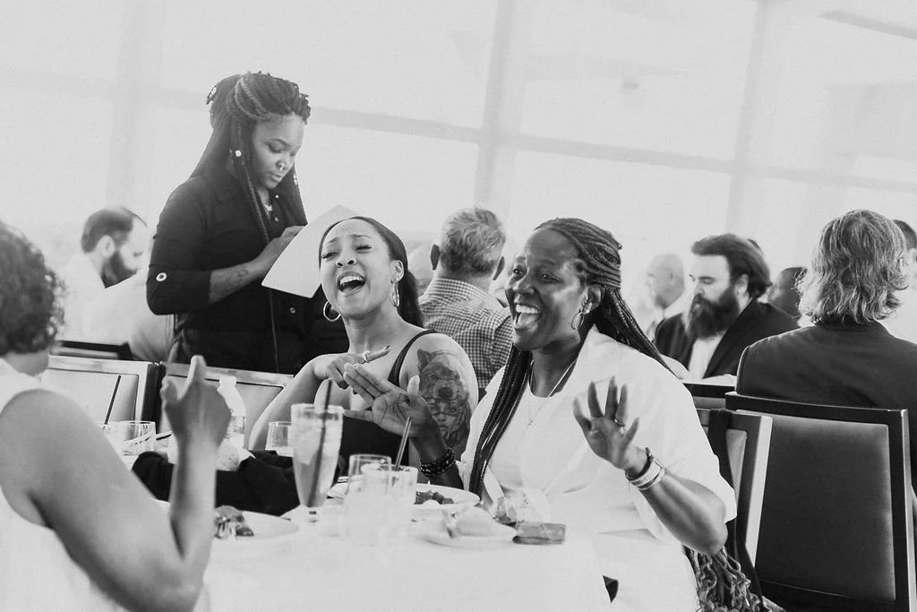 People sitting at a round table in a restaurant or banquet hall, laughing and engaging in conversation, with a waitress taking an order in the background.