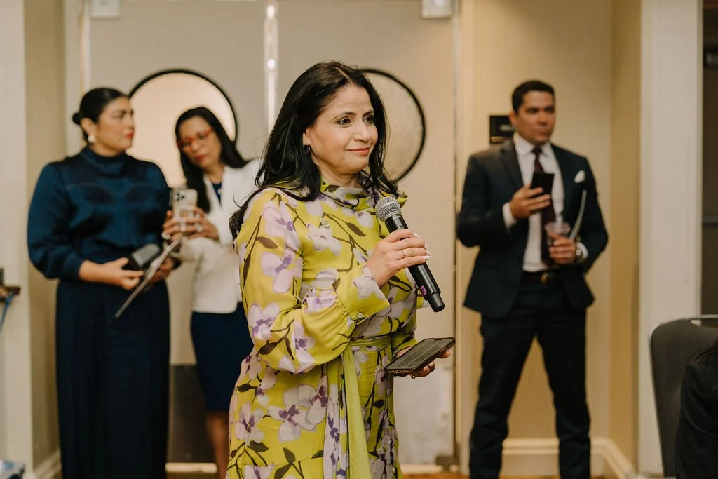 Woman in yellow floral dress speaking into a microphone at a formal event, with three people in the background, two women holding devices and a man in a suit.