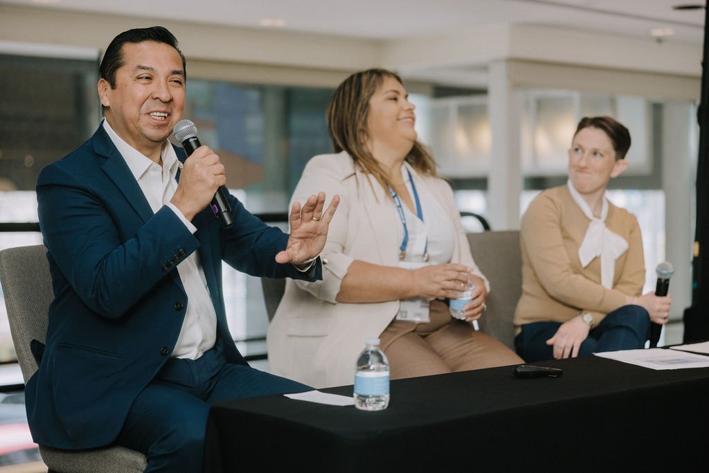 Three people sitting at a table with microphones, participating in a panel discussion or conference. One man in a blue suit is speaking into a microphone, while two women listen attentively, one holding a water bottle.