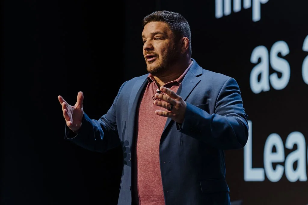A man speaking on stage in front of a screen, wearing a blue suit jacket and a maroon shirt.