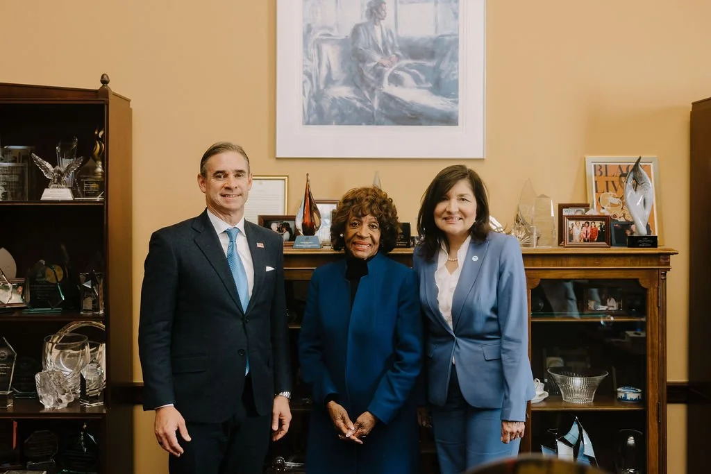 Three people standing in a decorated room, two women and one man, all dressed in business attire, smiling at the camera, with shelves and artwork in the background.