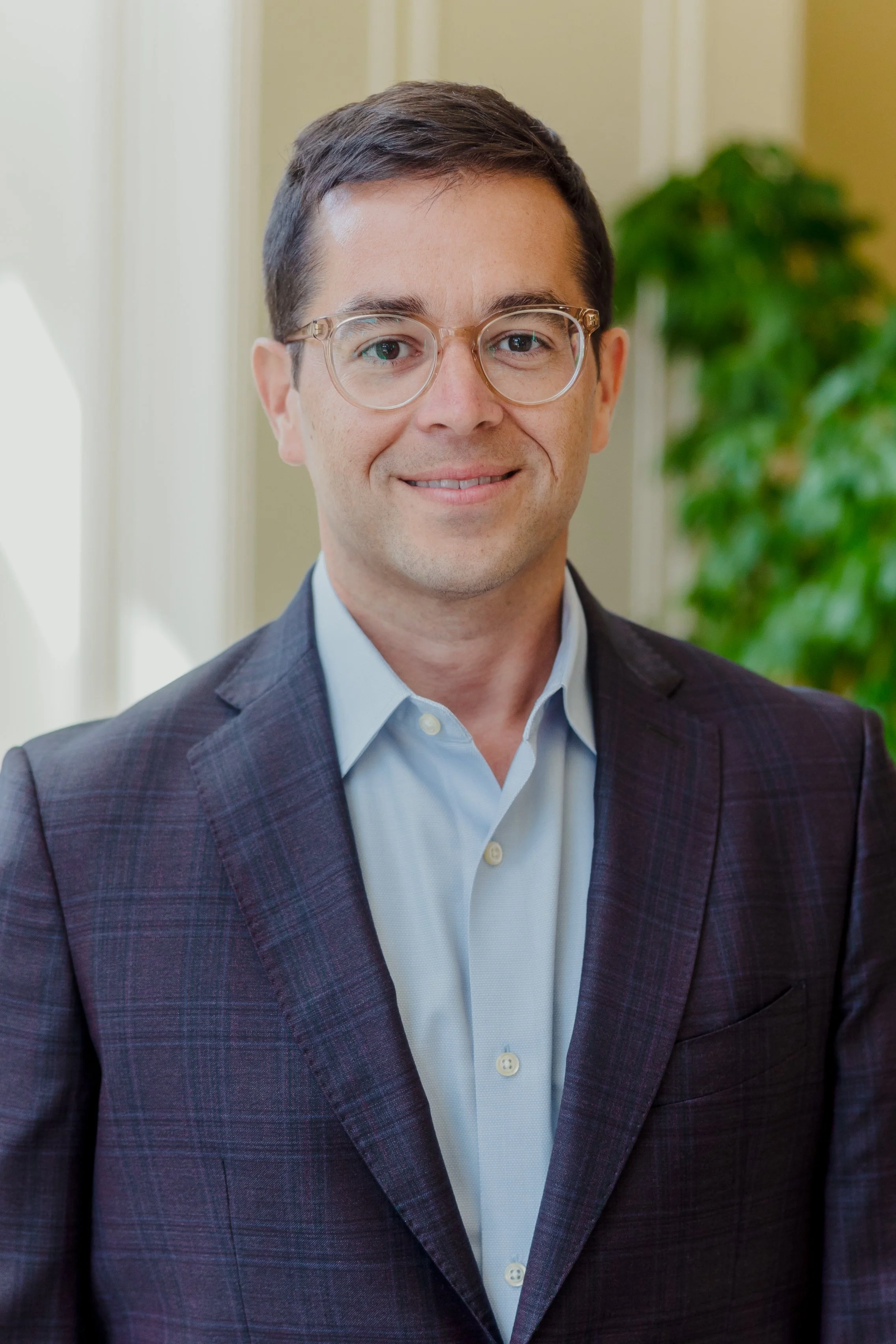 Portrait of a man with short dark hair, glasses, wearing a dark plaid suit jacket and light blue shirt, smiling, standing indoors with green plants in the background.