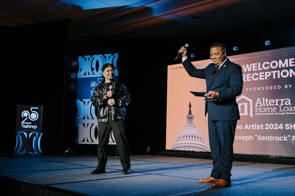 Two men speaking on stage at an event, one holding a microphone and the other holding a tablet, with a large screen behind them displaying the U.S. Capitol building and event information.