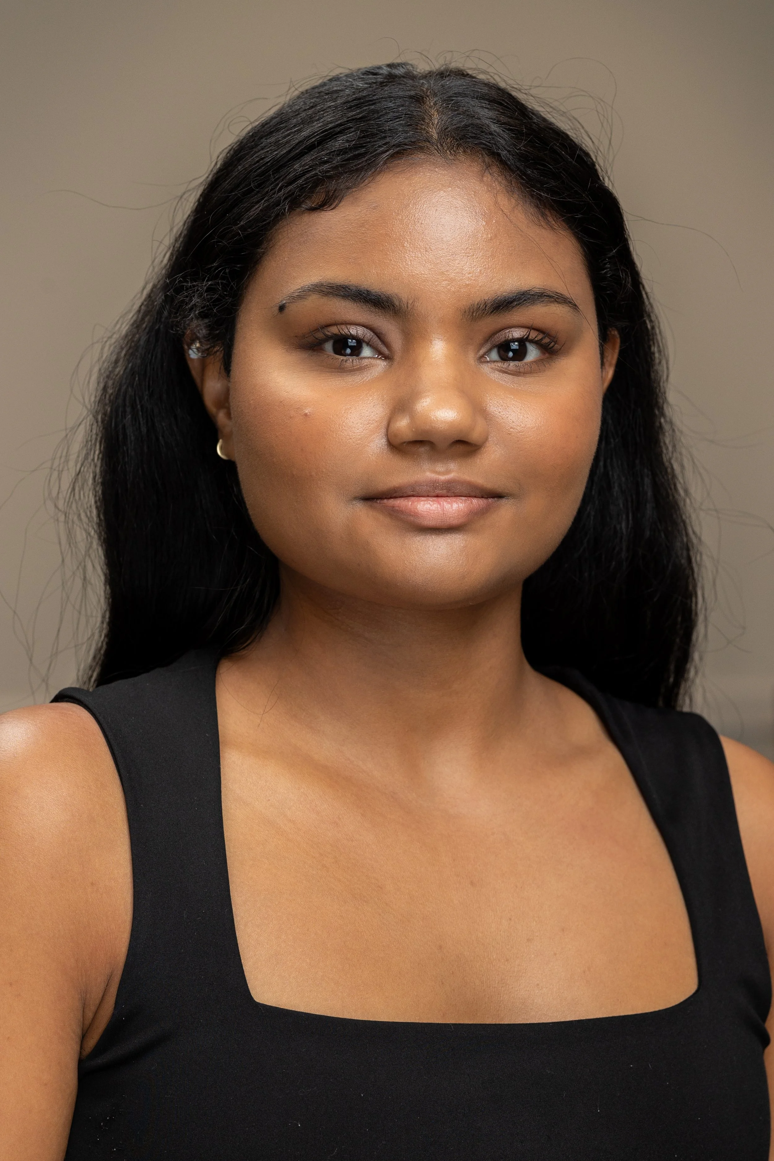 Portrait of a young woman with dark hair, wearing a black sleeveless top, standing against a plain background.