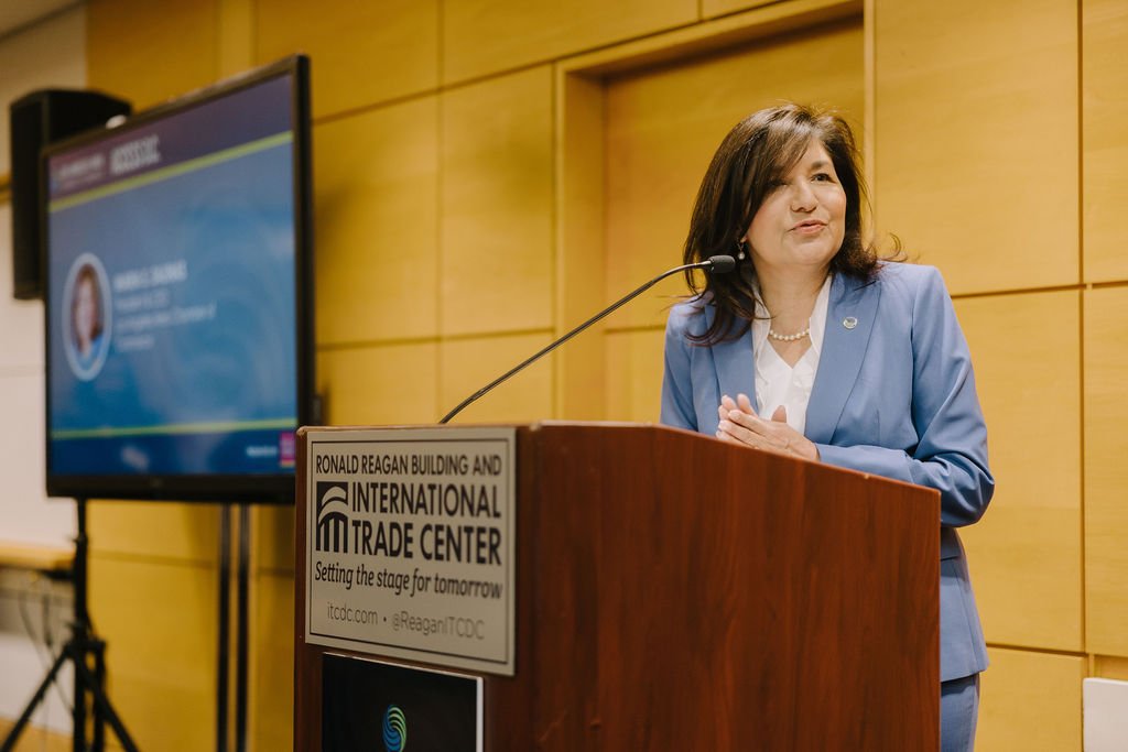 A woman in a blue blazer speaking at a podium in a conference room at the Ronald Reagan Building and International Trade Center.