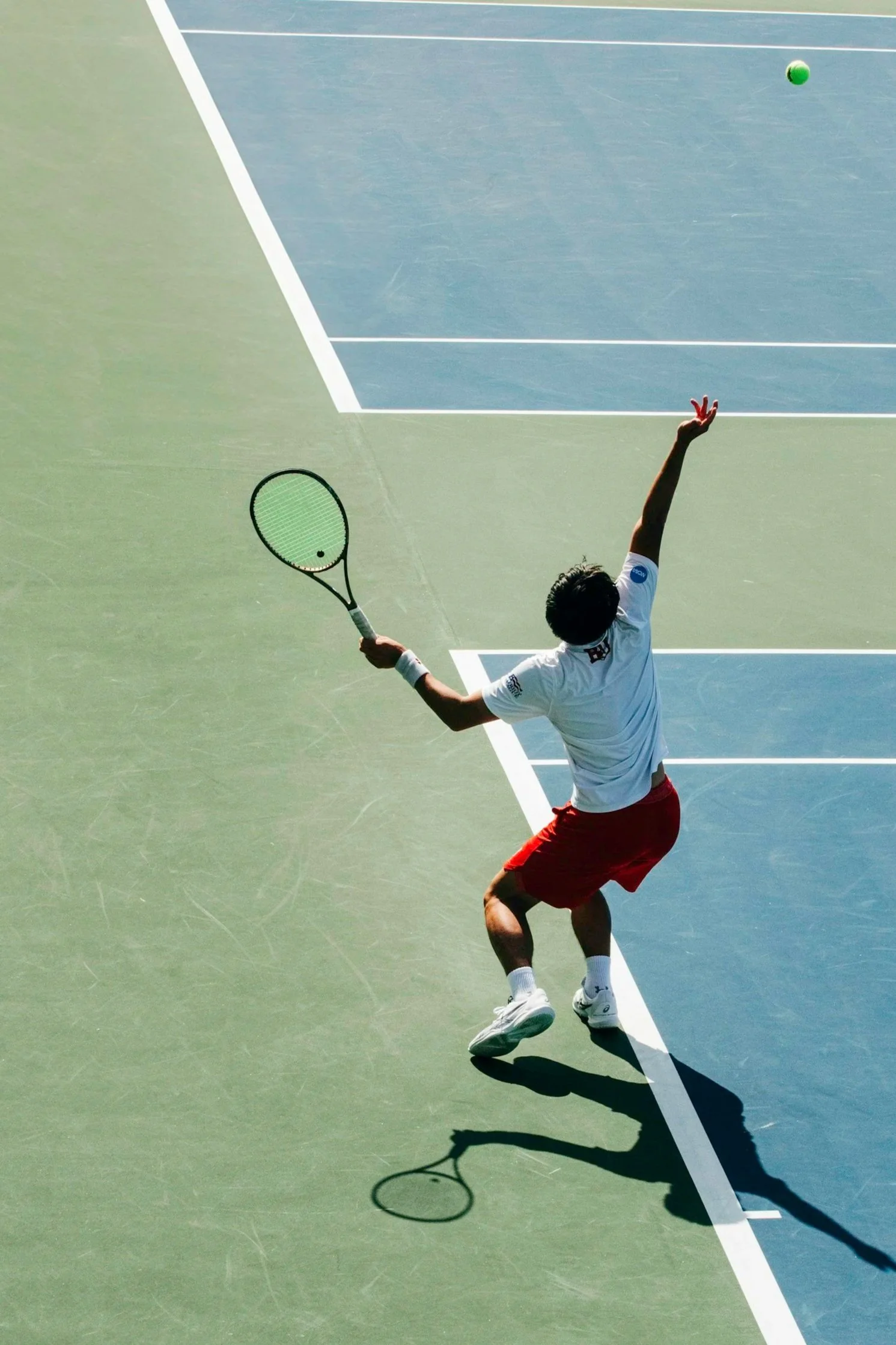 A tennis player in a white shirt and red shorts reaching back with his right arm while holding a tennis racket, on a tennis court with a green and blue surface, preparing to hit a green tennis ball.