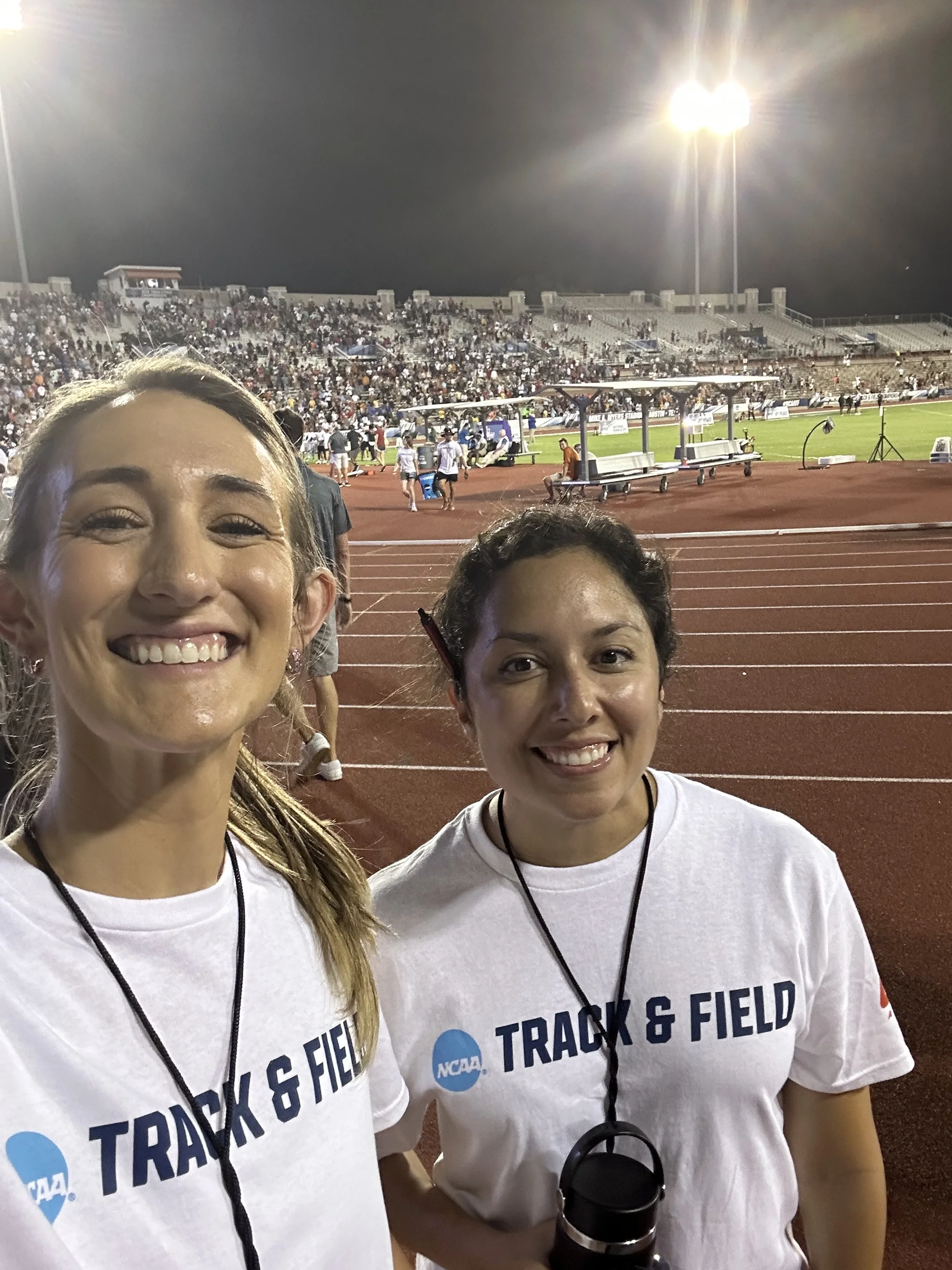 Two women smiling at a track and field event at night, wearing white T-shirts with 'NCAA Track & Field' printed on them, on a running track with a crowded stadium of spectators in the background.