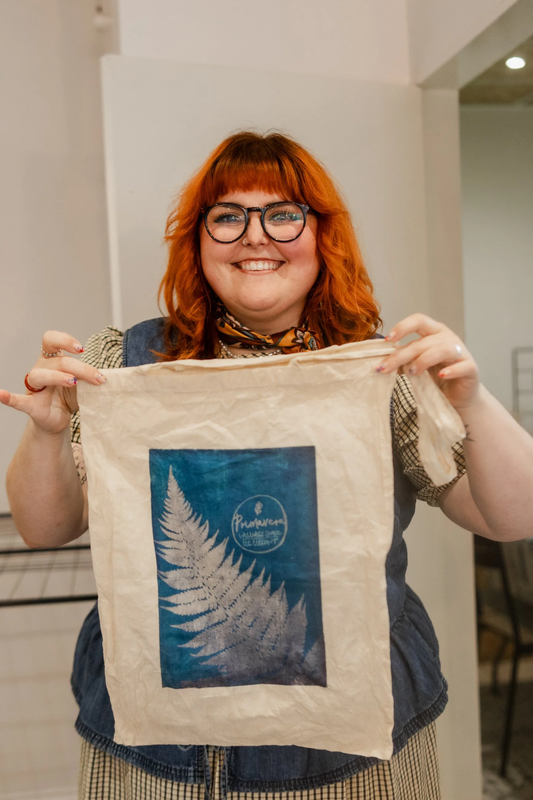 A woman with red hair, glasses, and a cheerful expression holding up a beige tote bag with a blue and white fern design and logo.