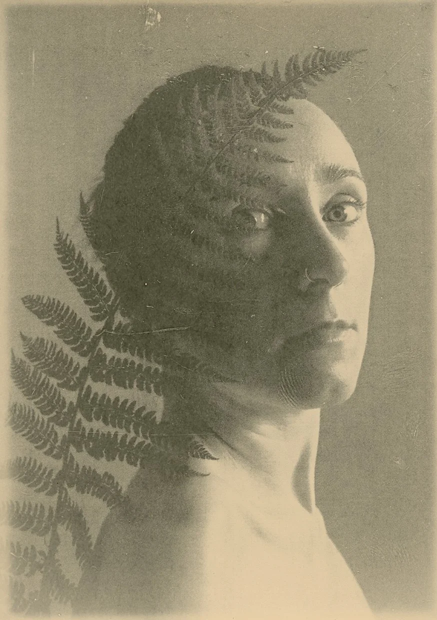 A black-and-white photographic portrait of a woman with her face in profile, partially covered by fern leaves, with light illuminating the right side of her face.