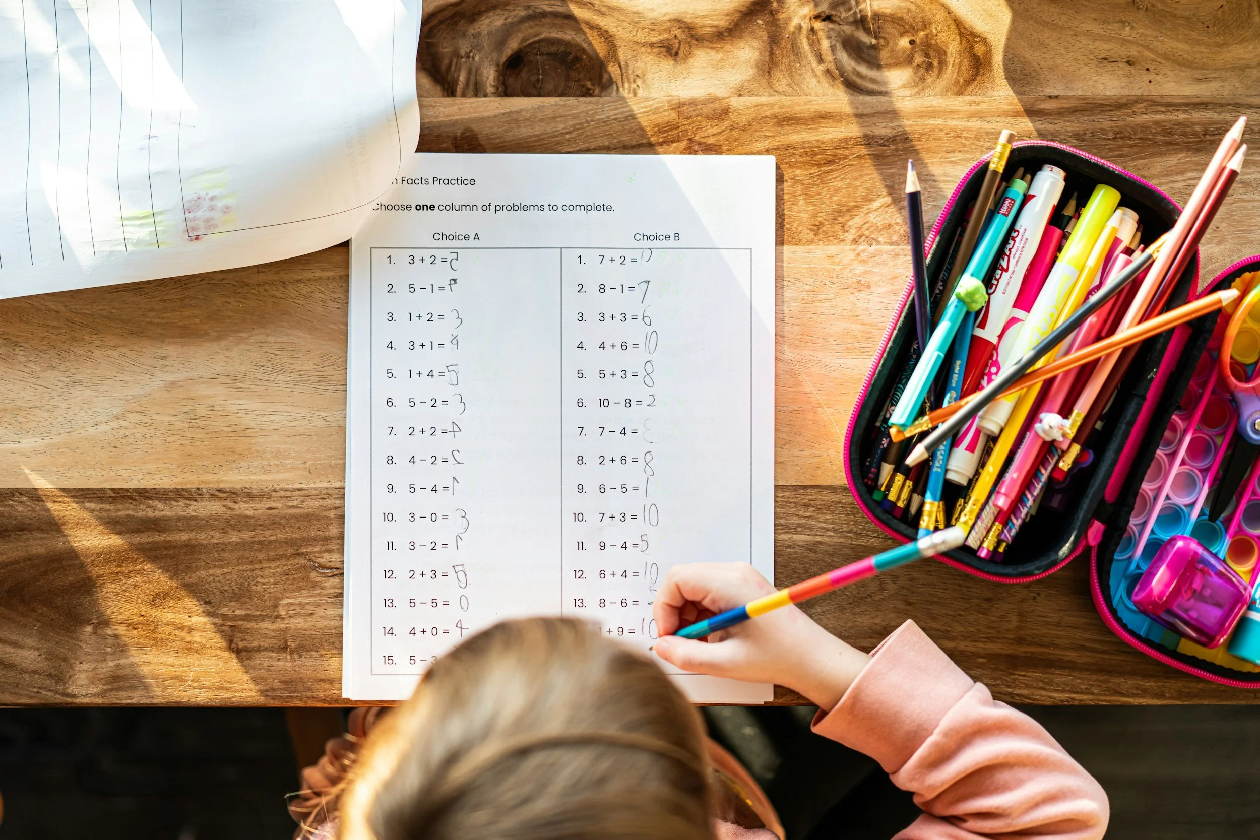 Child completing math worksheet with colorful pens and markers on a wooden table.
