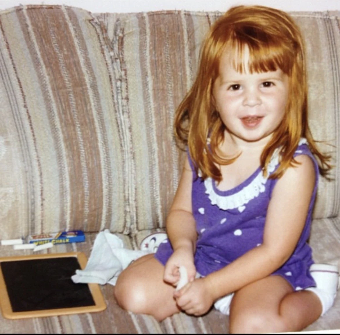 A young girl with red hair, wearing a purple polka-dot dress, sitting on a striped beige and brown couch, smiling at the camera while holding a piece of chalk, with a small black chalkboard, tissues, and a box of crayons nearby.