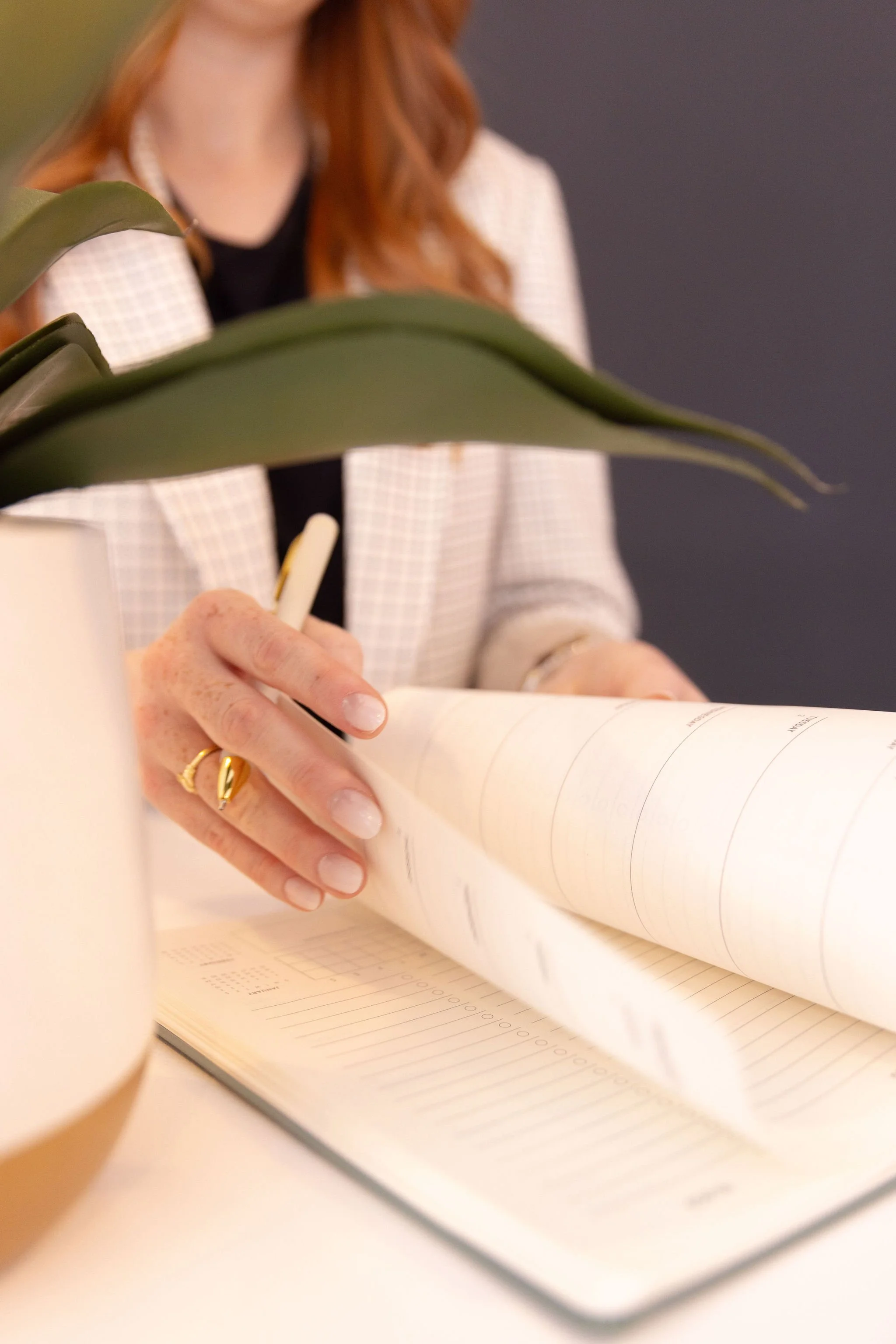 A person with red hair writing in a notebook with a pen, with a plant in the foreground and a cup in the lower left corner.