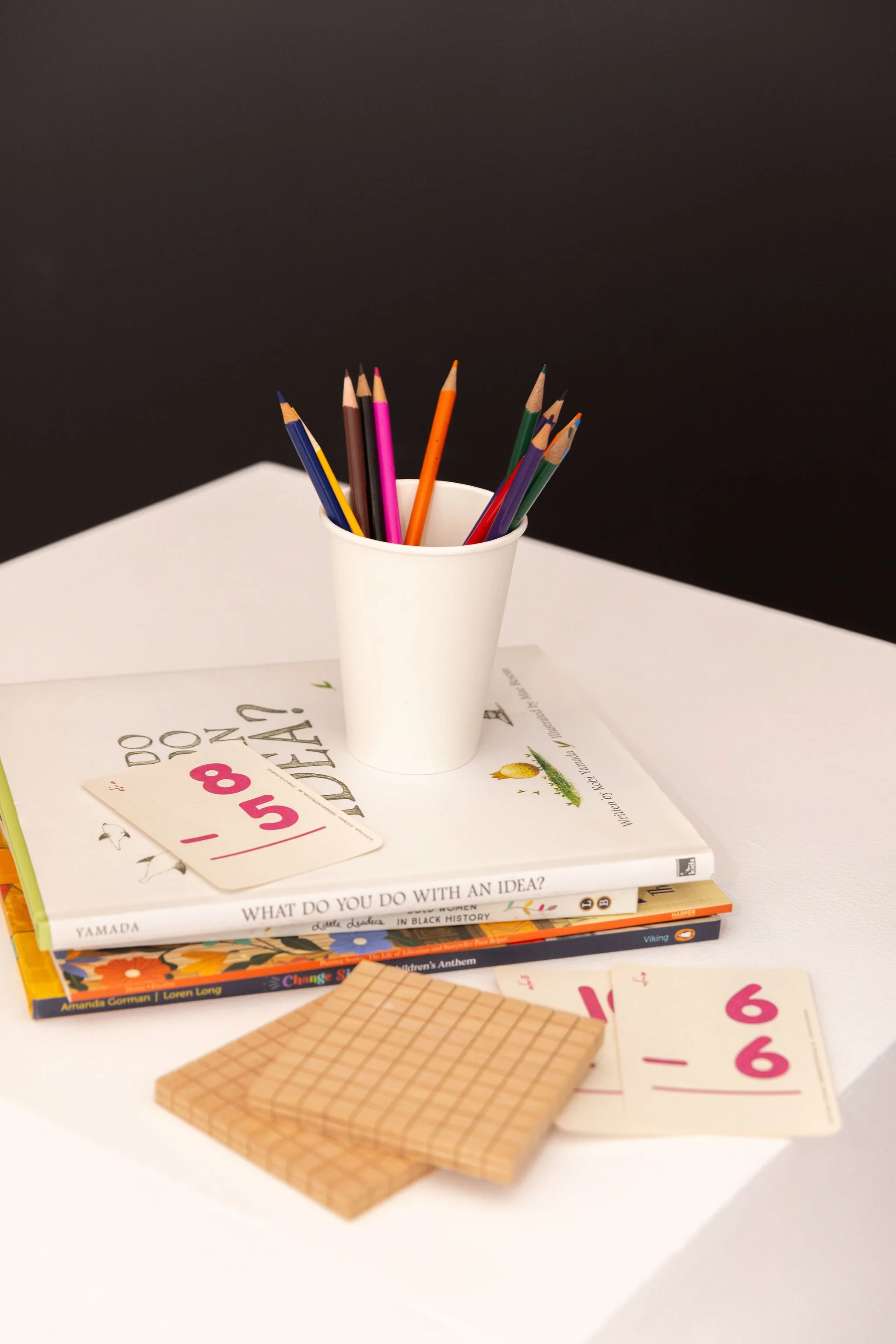 A white cup holding colored pencils on top of a stack of books and papers on a white surface with a dark background.