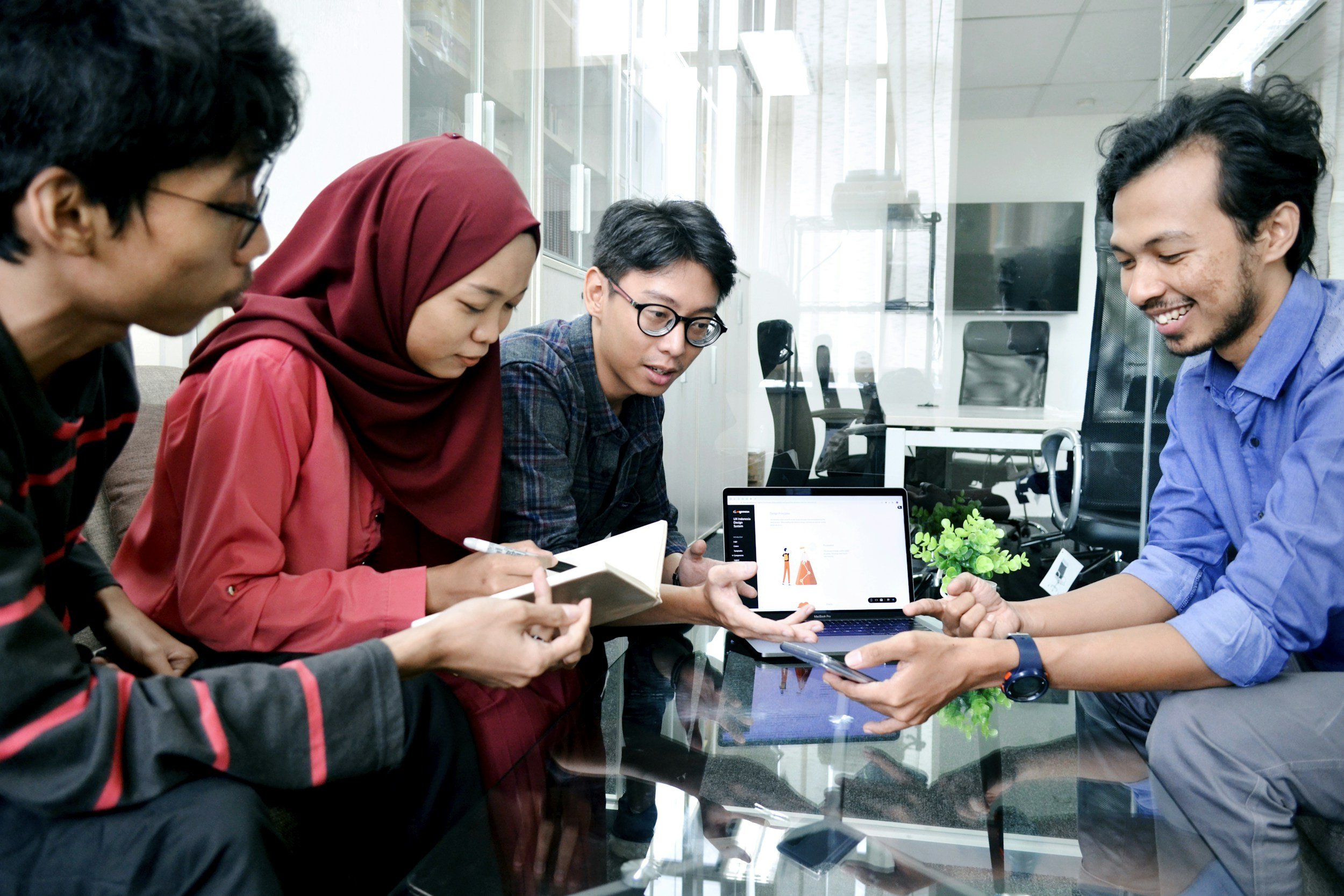 Group of five people gathered around a glass table, discussing and sharing ideas using a tablet and smartphone in an office.