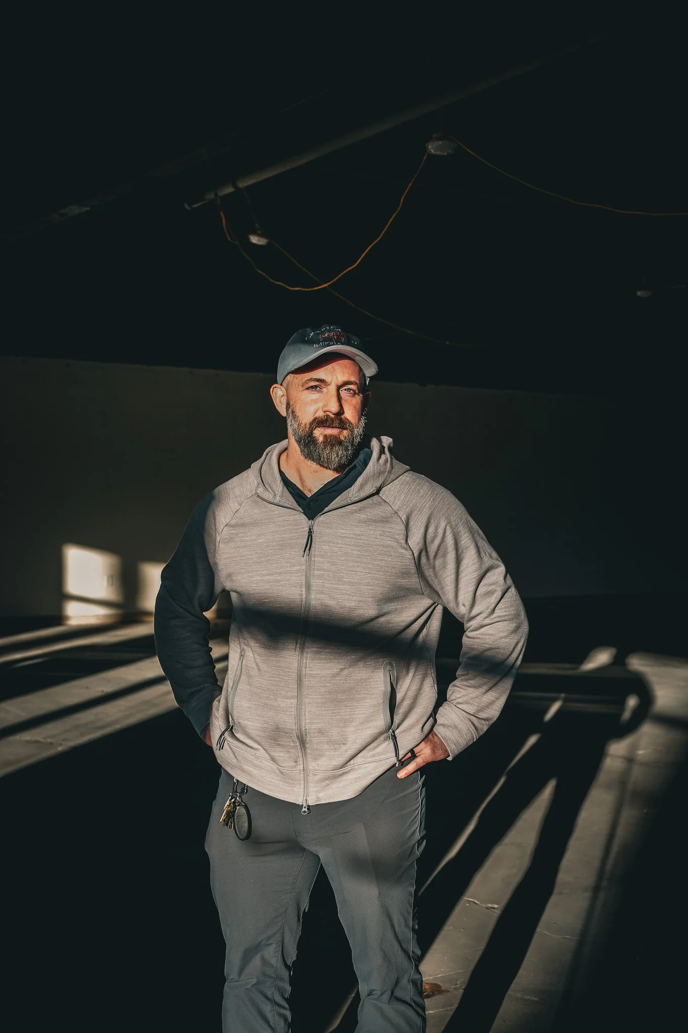 A bearded man wearing a gray hoodie and a baseball cap standing indoors in a room with dark walls, with sunlight casting shadows on the floor and wall.