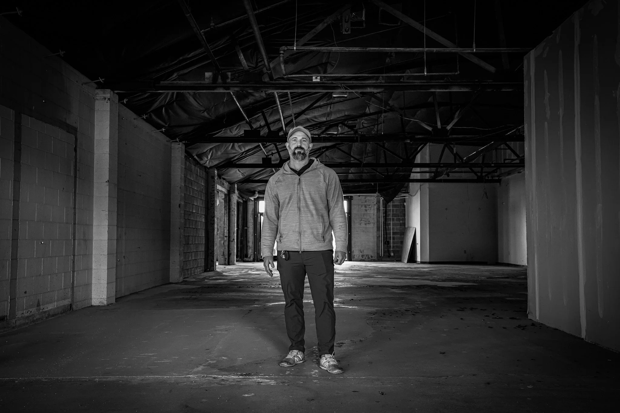 A man standing alone in an unfinished, empty building or warehouse with exposed beams and concrete floors, in black and white.