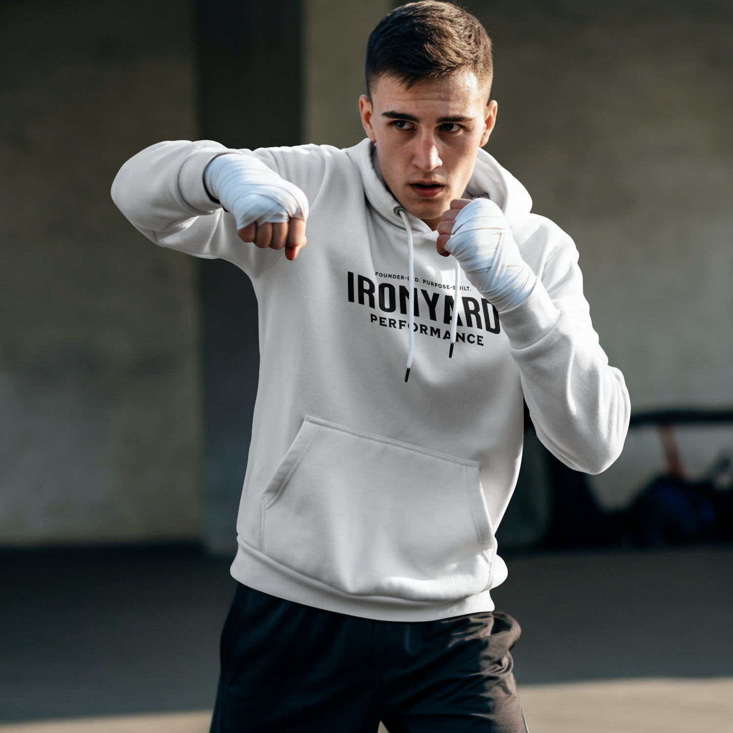 Young man wearing a white hoodie with 'IronYard Performance' text, practicing boxing with wrapped hands in a gym.