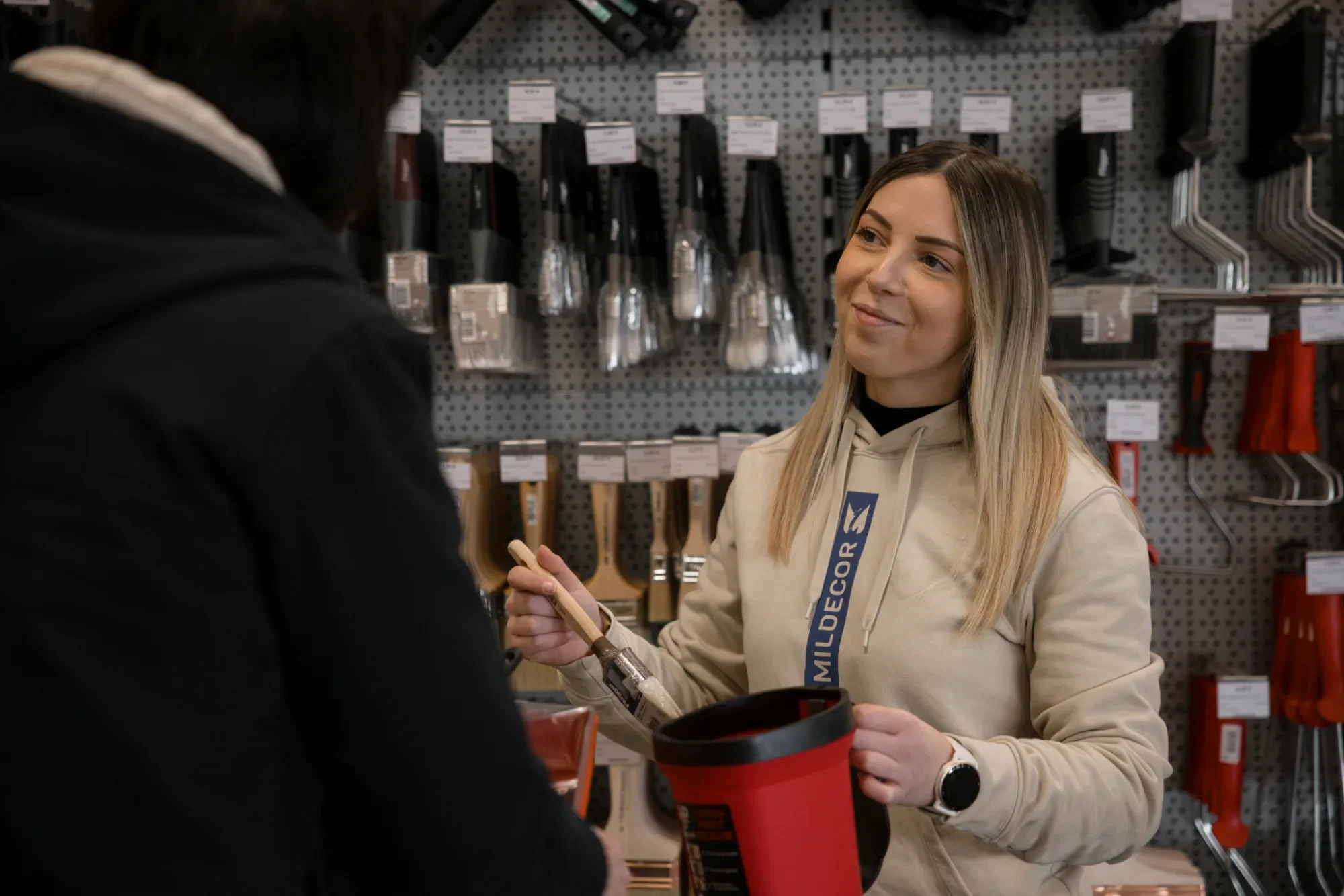 Une jeune femme, souriante, en train de conseiller un client dans un magasin de bricolage, tenant un pinceau et un pot de peinture, avec des outils suspendus en arrière-plan.