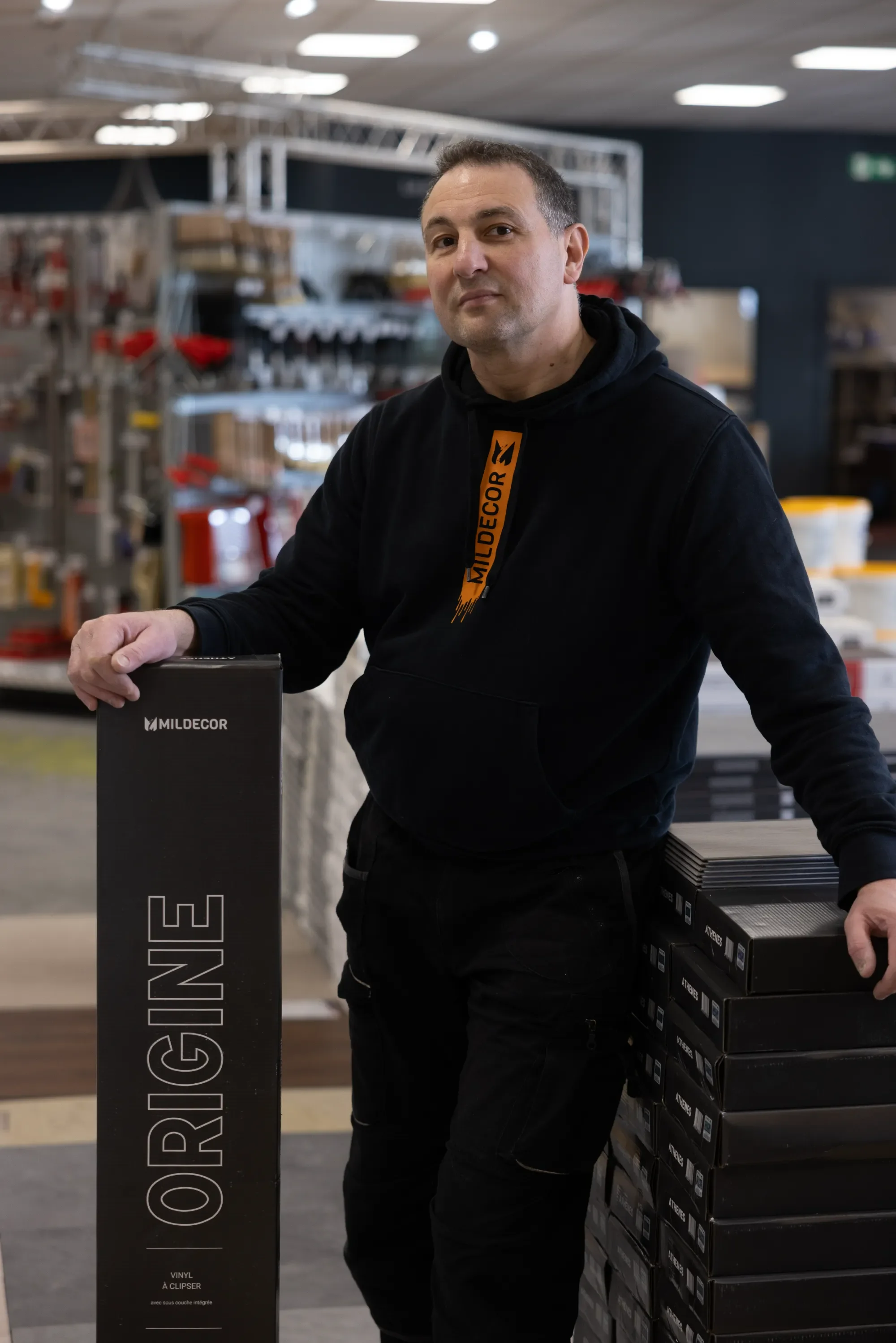 Homme dans un magasin de bricolage, portant un sweat à capuche noir avec le logo 'MILDECOR', tenant un emballage noir marqué 'ORIGINE' et entouré d'étagères avec des outils et des matériaux de construction.