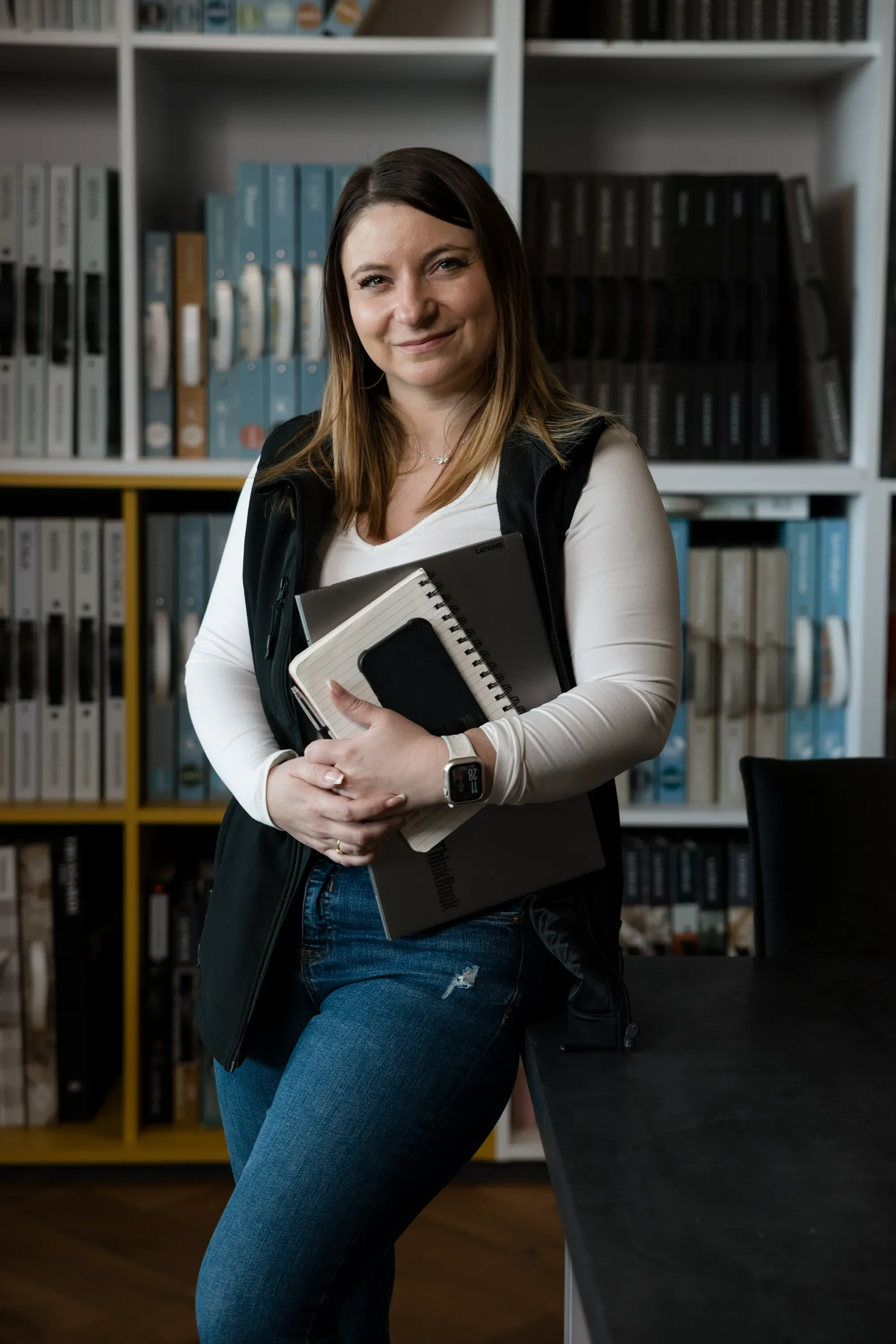 Femme souriante tenant des ordinateurs portables et des cahiers, portant une montre connectée, en bibliothèque ou bureau.