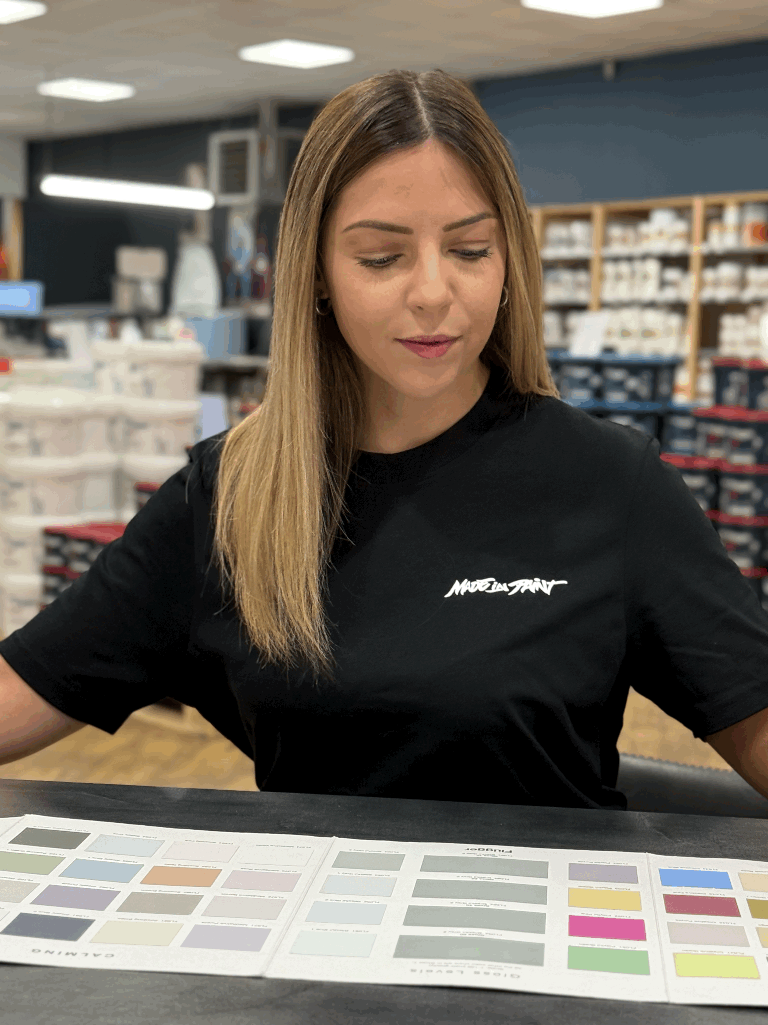 Une femme aux cheveux lisses, portant un t-shirt noir avec un logo blanc, assise à une table avec un grand échantillon de couleurs, dans un magasin ou une surface de vente.