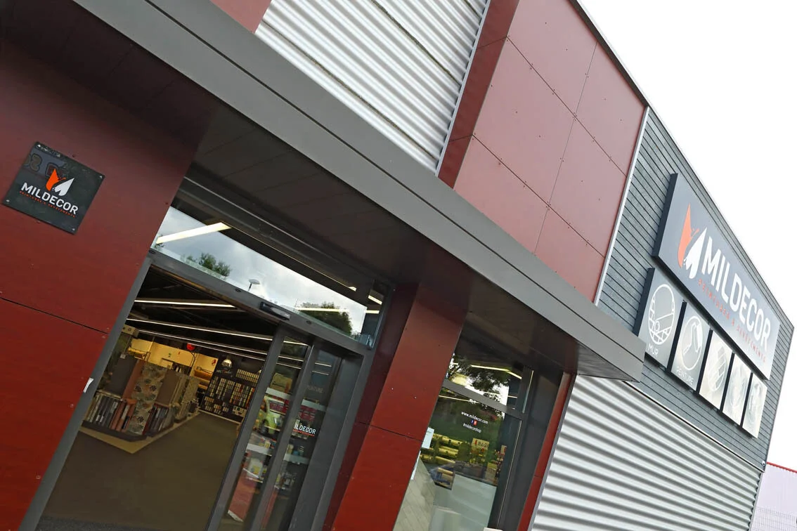 Une façade d'un magasin de matériaux de construction nommé Mildecor avec un panneau illustrant différents types de produits comme peinture, revêtement mural et autres.