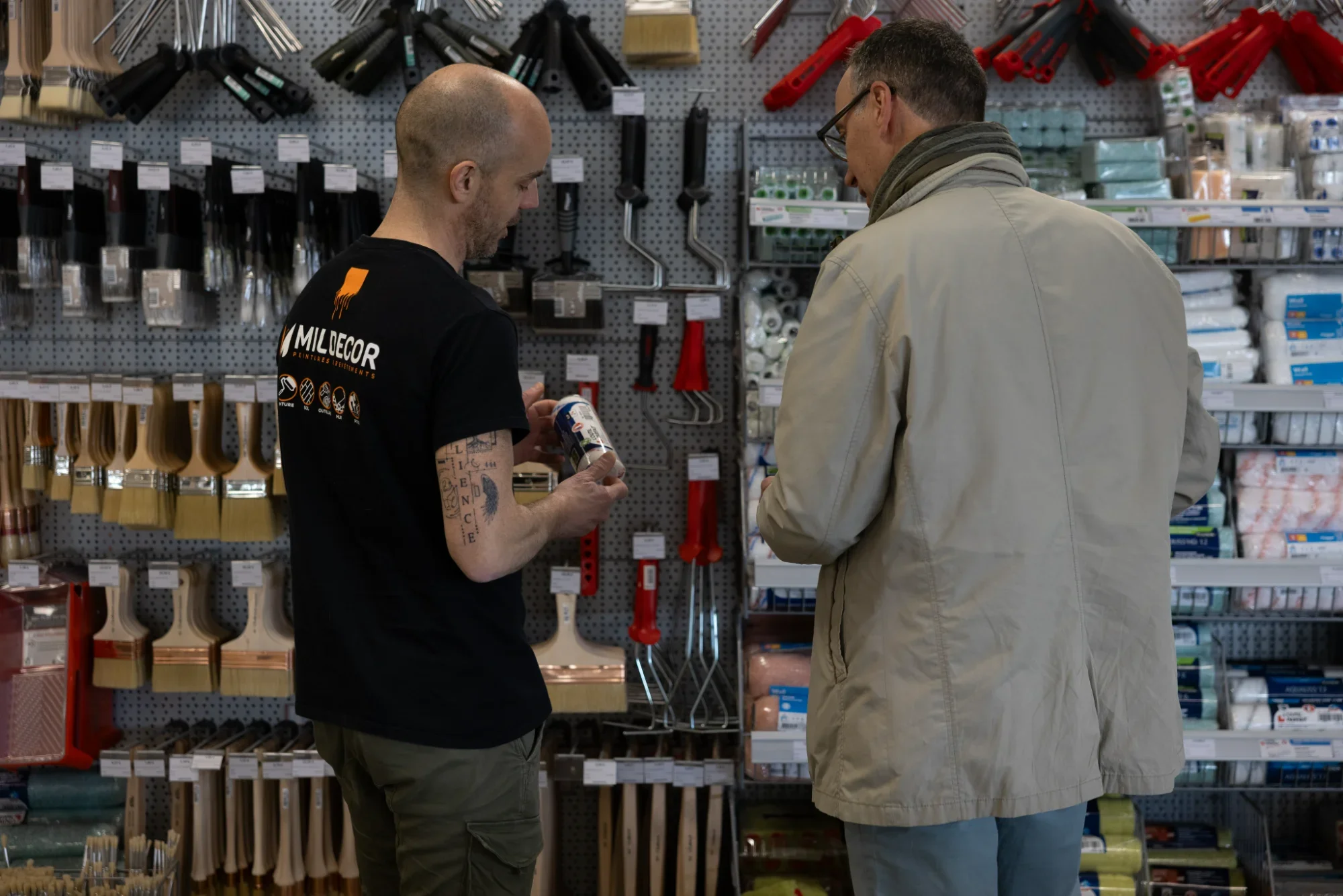 Deux hommes dans un magasin de bricolage, l'un portant un t-shirt noir avec le logo 'MILDECOR' et l'autre vêtu d'une veste beige, discutent en regardant un produit. Le fond montre des étagères remplies d'outils et de fournitures de bricolage.