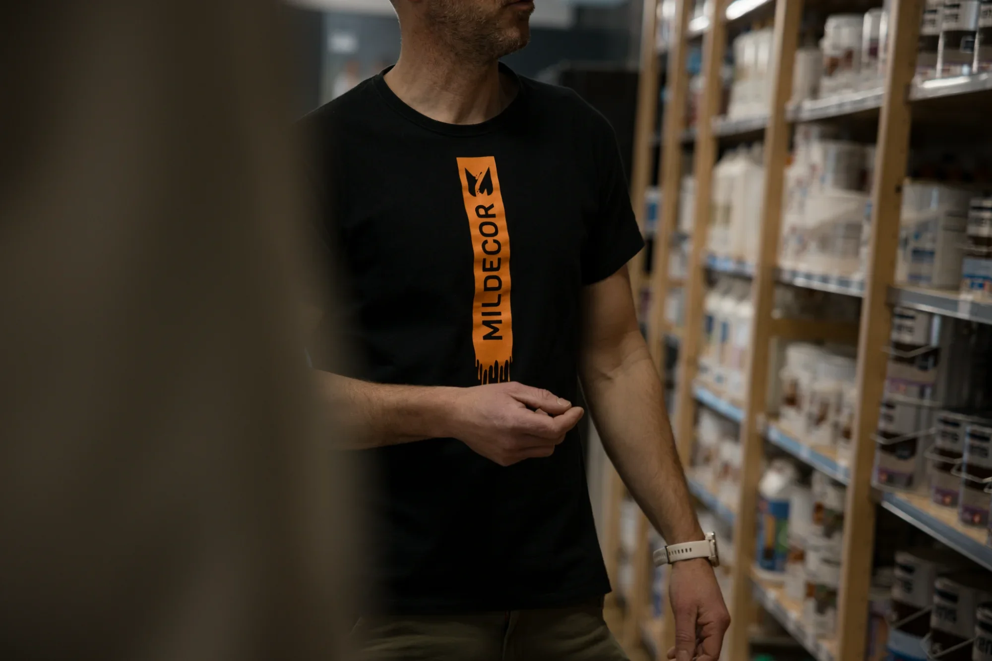 Une personne portant un t-shirt noir avec la inscription orange 'MILEDECORE S' dans une bibliothèque ou un magasin avec des étagères remplies de produits.