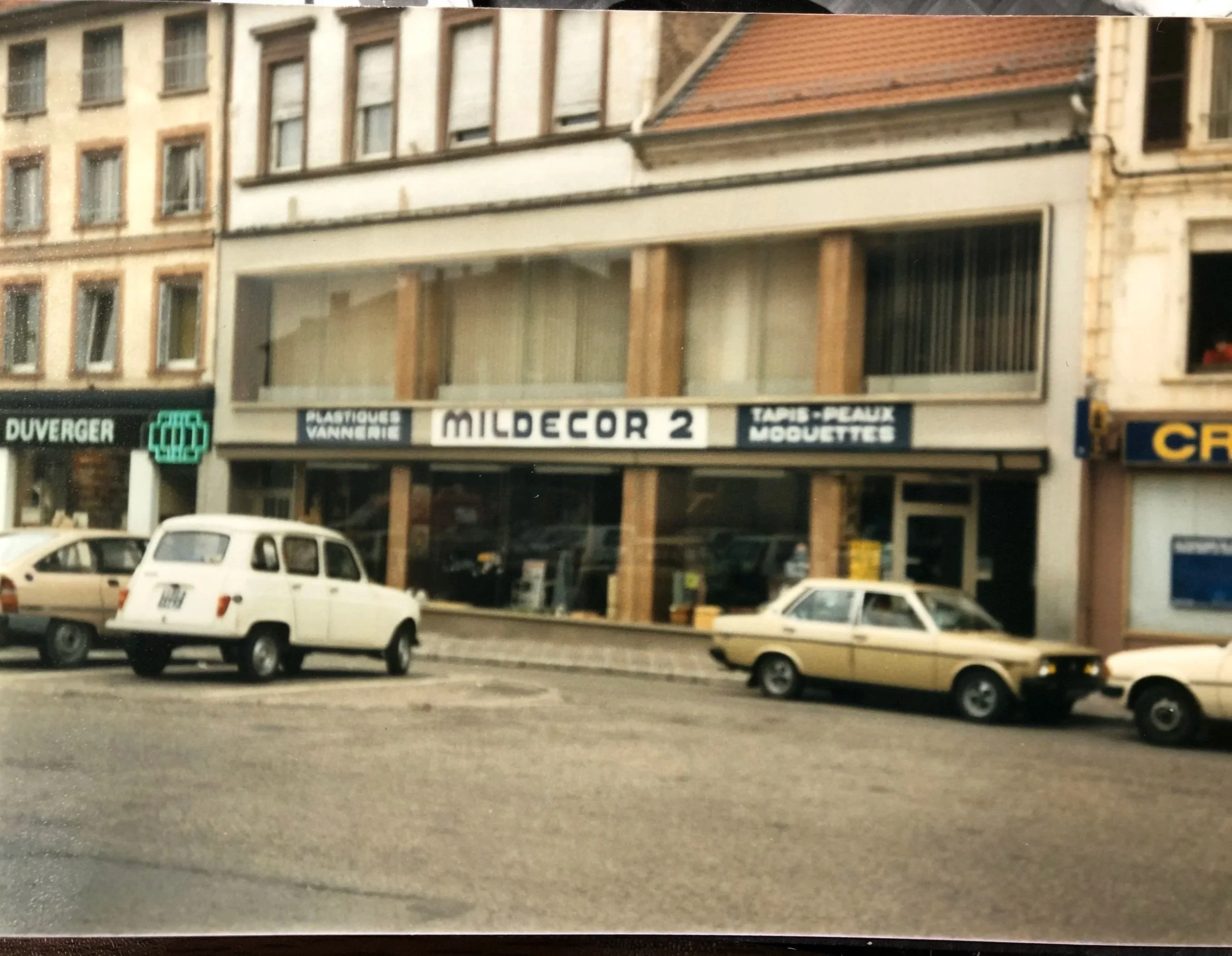 Une vue de la façade d'un immeuble résidentiel avec plusieurs fenêtres. Au rez-de-chaussée, il y a des commerces, dont une boutique appelée 'Mildecor 2' offrant des panneaux en pvc, des tapis et moquettes. En avant, il y a une rangée de voitures stationnées.