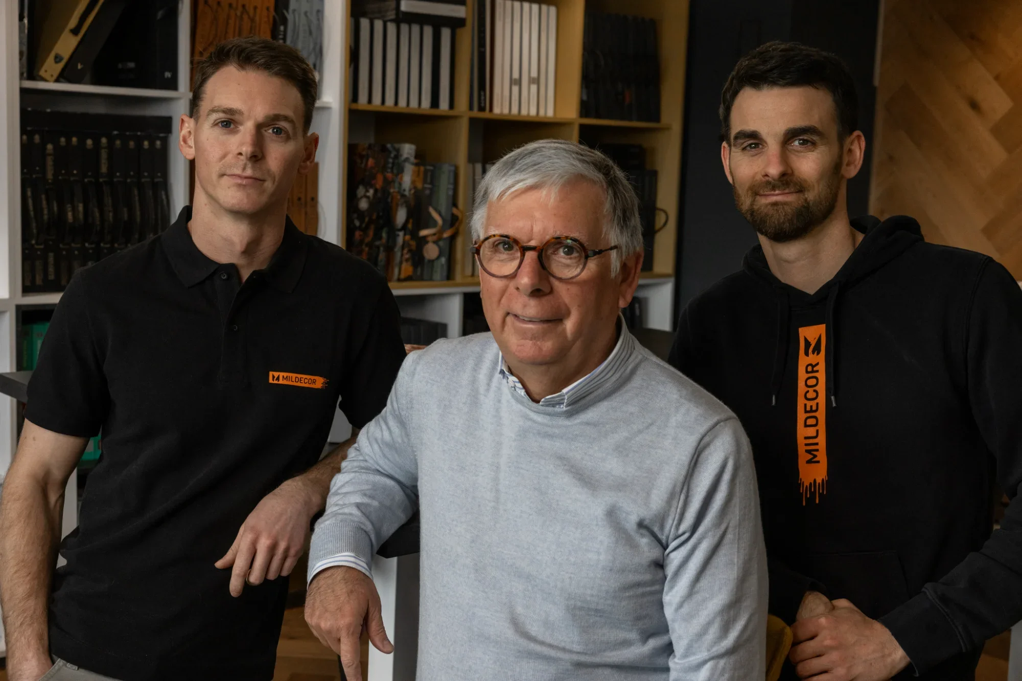 Trois hommes posent ensemble dans une bibliothèque, un homme âgé au centre portant des lunettes, flanqué de deux jeunes hommes en vêtements noirs avec des logos 'Mildecors'.