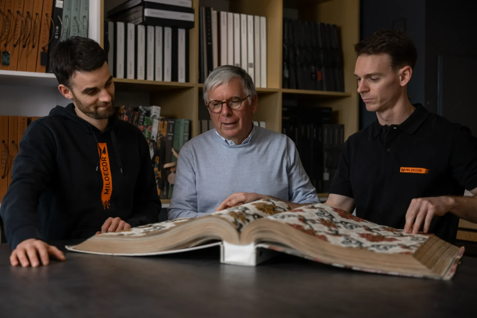 Trois personnes examinant un grand livre ouvert en bibliothèque avec des étagères remplies de fichiers et livres en arrière-plan.