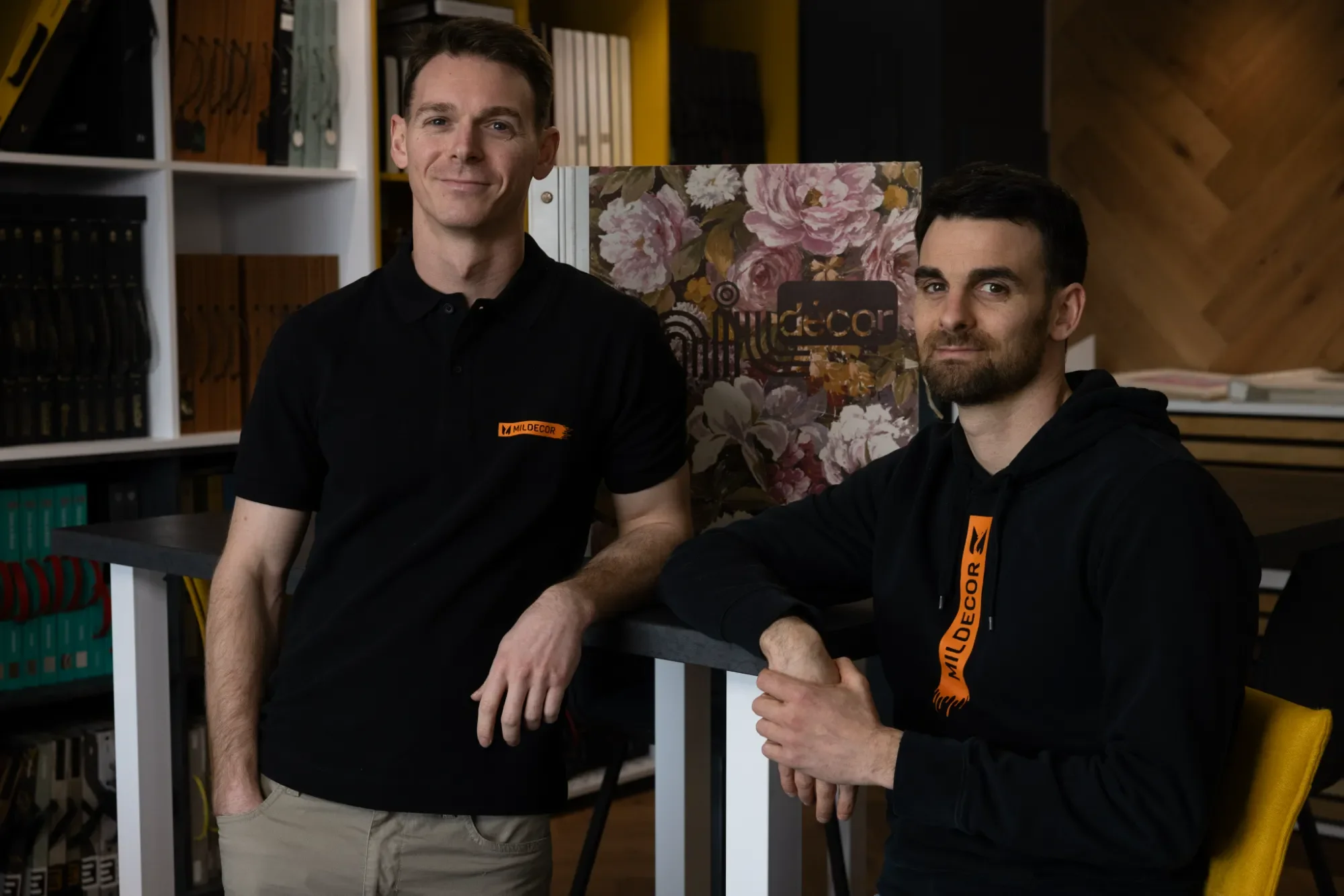Deux hommes posent dans un espace de bureau ou de magasin, portant des vêtements noirs avec un logo orange de Milfdecors. L'un se tient debout, l'autre est assis. Il y a des étagères remplies de livres ou de fichiers en arrière-plan, et un panneau dé
