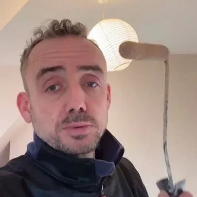 Un homme avec des cheveux courts et une barbe, portant une veste noire, se tient dans une pièce avec un lampadaire en papier en arrière-plan, tenant un outil avec un fil.